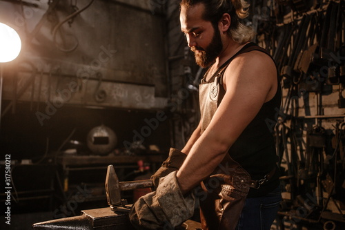 Portrait of a brutal muscular blacksmith standing in a dark workshop forging iron