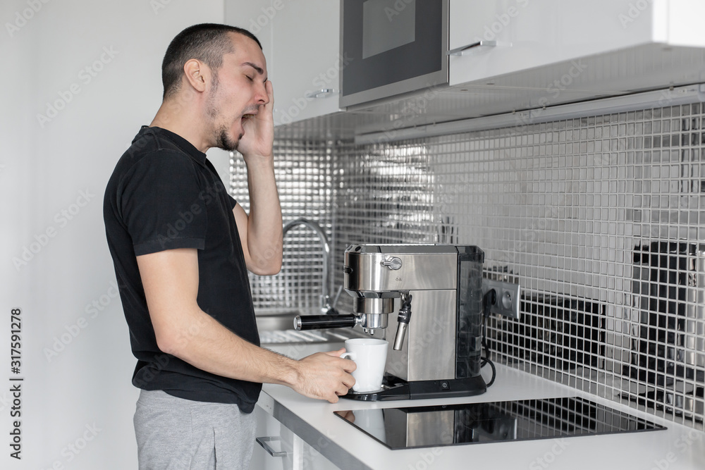 Sleepy guy in the morning in the kitchen preparing coffee in a coffee machine.