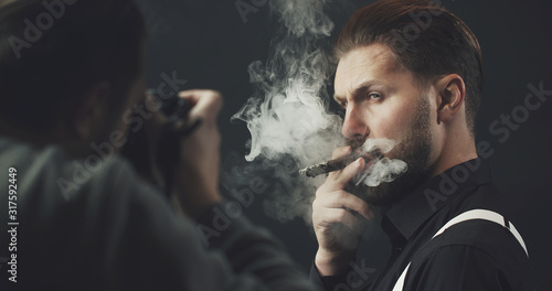 Photographer with camera shooting adult man smoking cigar over dark background, cropped shot