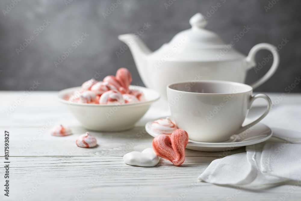 Romantic breakfast for a Valentines Day with tea and meringue cookies in the shape of hearts and candys. White tea-set stand on a wooden table with a concrete background. Close up still life. Romantic breakfast for a Valentines Day with tea and meringue cookies in the shape of hearts and candys. White tea-set stand on a wooden table with a concrete background. Close up still life.