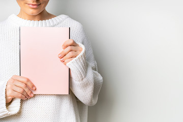 Close view of woman in white woolen sweater holding a book with empty pink cover in hands. Free space for your mock up of reading book concept background.