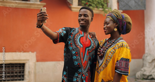 Принти на полотні African young stylish and attractive couple in traditional outfits standing together outside and posing to the smartphone camera while taking selfie photo
