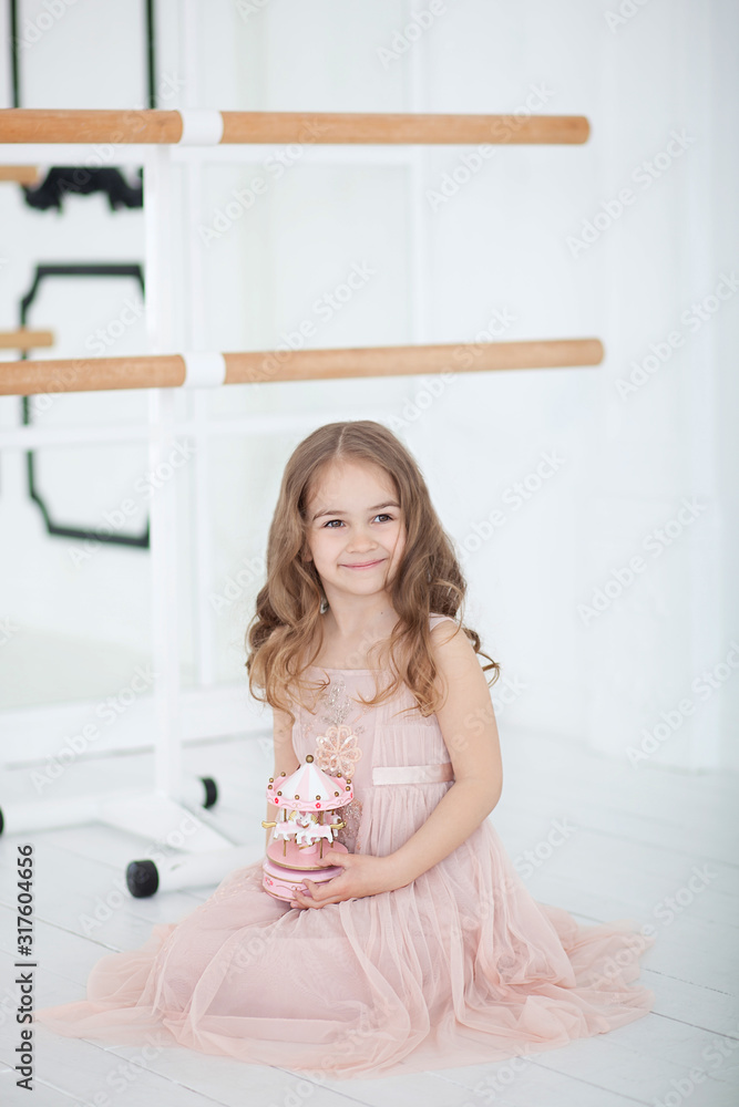 A little ballerina in a dress sits in a dance class on the floor. Little girl holding a musical toy carousel. The child receives a gift. Vintage musical carousel toy. indoors ballet hall class room