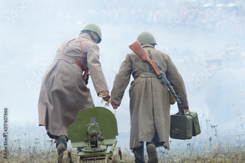 Soldiers of the Soviet army in helmets with weapons behind their backs roll a green machine gun of the Maxim system across the field in smoke. Reconstructing World War II battles photo
