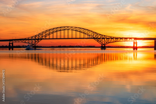  Steel tied arch bridge spanning a bay with crystal clear reflections in the water at sunset. Fire Island Inlet Bridge, part of the Robert Moses Causeway on Long Island New York. 