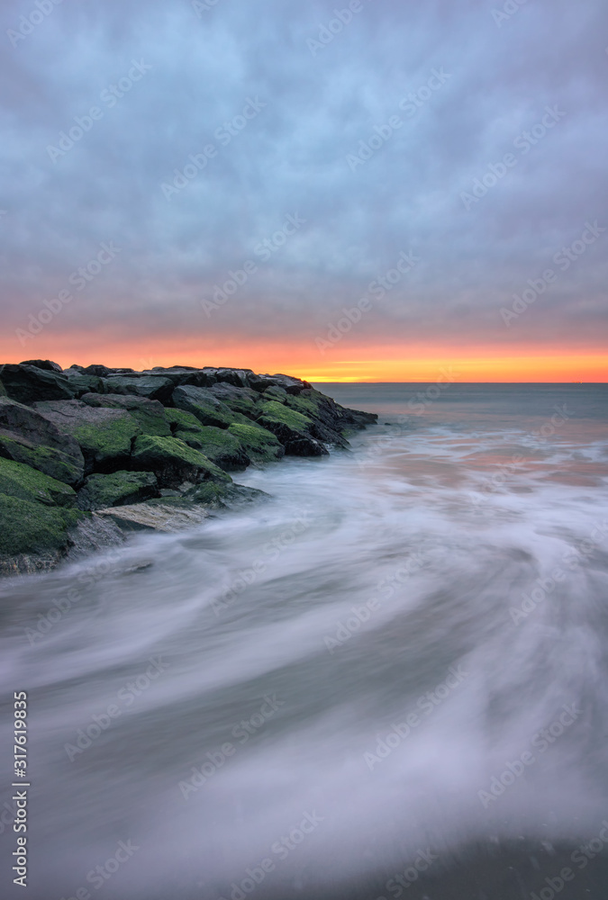 Fototapeta premium Dark ominous storm clouds and a sliver of golden light over a stone jetty on a beach, with swirling waves in motion. Long Beach, New York. 
