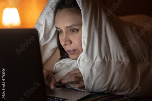 Woman looks with interest at a laptop, hiding under a blanket, in bed, at night by the light of a lamp. photo