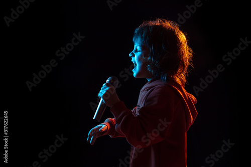 Singing like celebrity  rockstar. Caucasian boy s portrait on dark studio background in neon light. Beautiful curly model. Concept of human emotions  facial expression  sales  ad  music  hobby  dream.