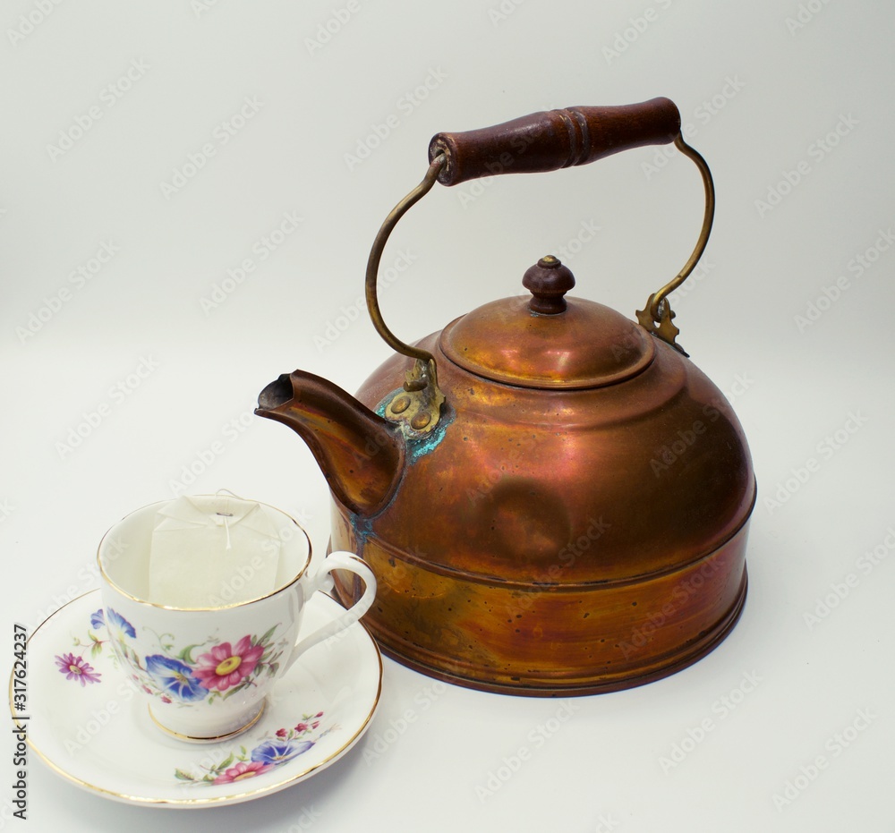 Close up on old copper tea kettle next to flower themed tea cup and saucer. Close up on old copper tea kettle next to flower themed tea cup and saucer.