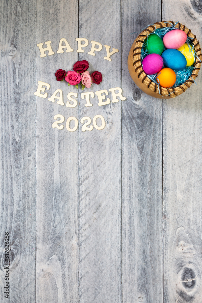 Easter eggs in a basket with Happy Easter 2020 and flowers on a wood background with copy space