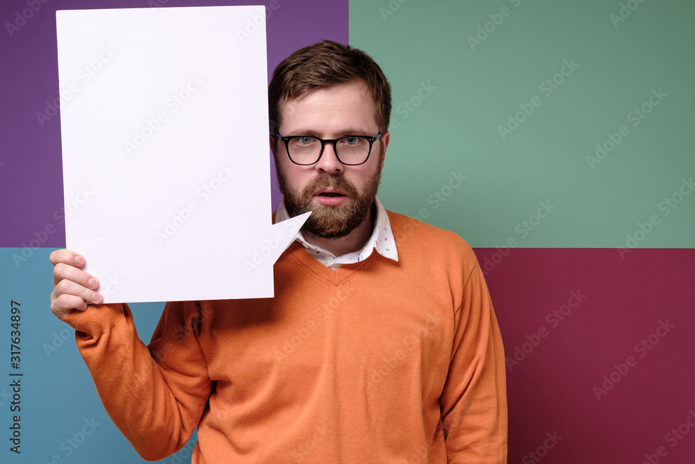 Bearded man with a strange expression on his face is holding a blank, white board, advertising space or announcement.