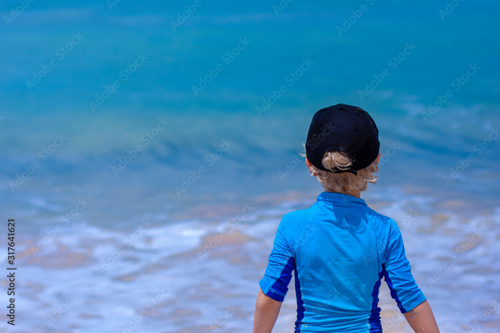 A boy in a black cap and blue swimming top looking to the ocean. Finger Bay, Australia. A boy in a black cap and blue swimming top looking to the ocean. Finger Bay, Australia.