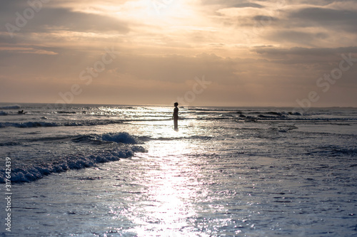 A silhouette of a young person stand in the water lit by the path of the rising sun on the ocean beach. Anna Bay, Australia