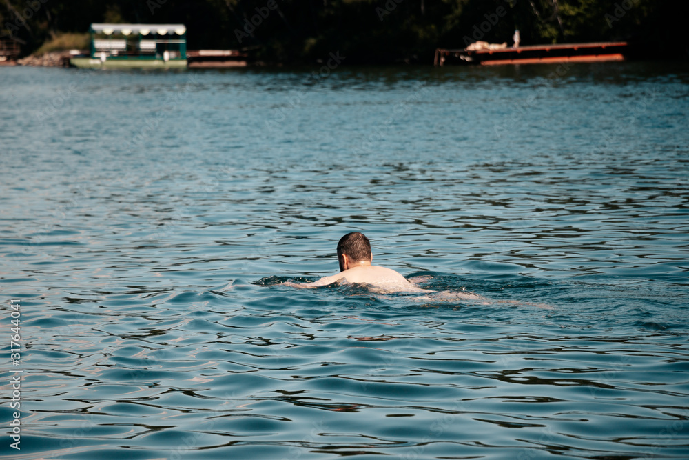 A man swims along the shore of the lake. The coast of the lake with boats. A man in the water. The blue expanse of the lake.