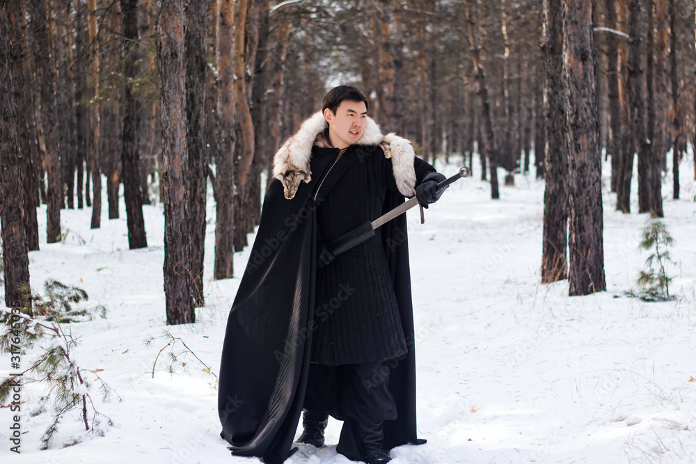 A warrior in black quilted clothes in a black cloak with a white fur collar and a long sword in his hands walks along the road in the forest.