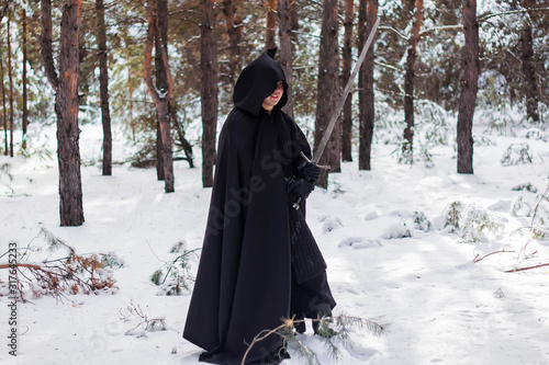A warrior in black quilted clothes, in a black cloak with a hood on his head and a long sword in his hands against the background of winter forest and snow.