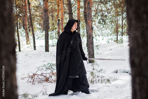 A warrior in black quilted clothes, in a black cloak with a hood on his head and a long sword in his hands against the background of winter forest and snow.