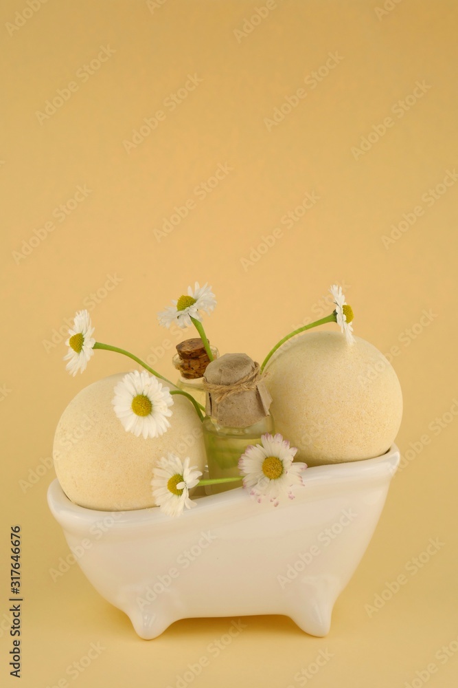 Bath bombs with chamomile extract.Organic natural body cosmetics. Two bath bomb balls and daisy flowers in a decorative white bath on a light yellow background.