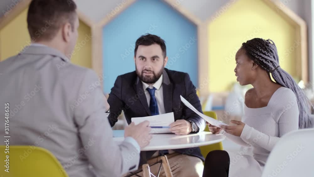 Three businessmen of different races at the negotiating table, study documents before signing a new contract. A young African American business woman speaks to her opponent through her human Three businessmen of different races at the negotiating table, study documents before signing a new contract. A young African American business woman speaks to her opponent through her human
