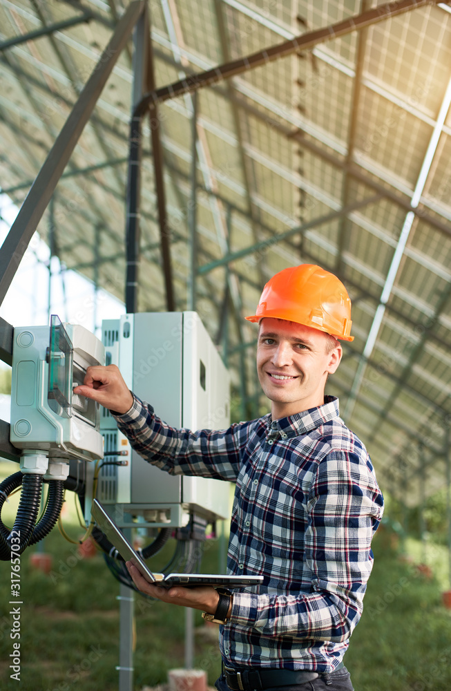 Smiling woker in helmet and with laptop at station pushing button at the electrical equipment on solar power station. Vertical shot. Science technology. Solar panels. Global warming. Green energy. Smiling woker in helmet and with laptop at station pushing button at the electrical equipment on solar power station. Vertical shot. Science technology. Solar panels. Global warming. Green energy.