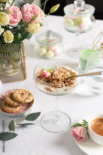 Breakfast concept, Sunday brunch in a restaurant. Light granola with Greek yogurt in a glass goblet, coffee, cereal cookies, sweets and flowers on a snow-white tablecloth.