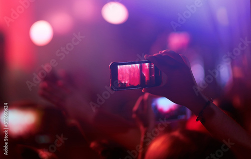 Hand with a phone records live music festival. People taking photographs with smart phone during a public music concert. Crowd raising their hands, dancing and enjoying great the concert.