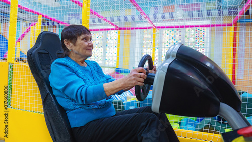 Satisfied aged lady smiling while sitting in car driving simulator attraction in children s amusement park