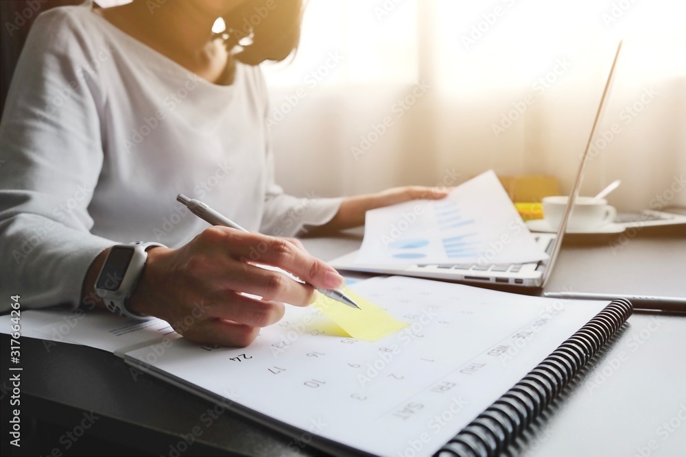 Asian woman is writing on yellow paper notes and put a warning on calendar. The other hand hold the report graph and has laptop on the front. Maybe preparing data for marketing report. window light Asian woman is writing on yellow paper notes and put a warning on calendar. The other hand hold the report graph and has laptop on the front. Maybe preparing data for marketing report. window light