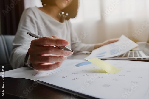 Asian woman is writing on yellow paper notes and put a warning on calendar. The other hand hold the report graph and has laptop on the front. Maybe preparing data for marketing report. window light