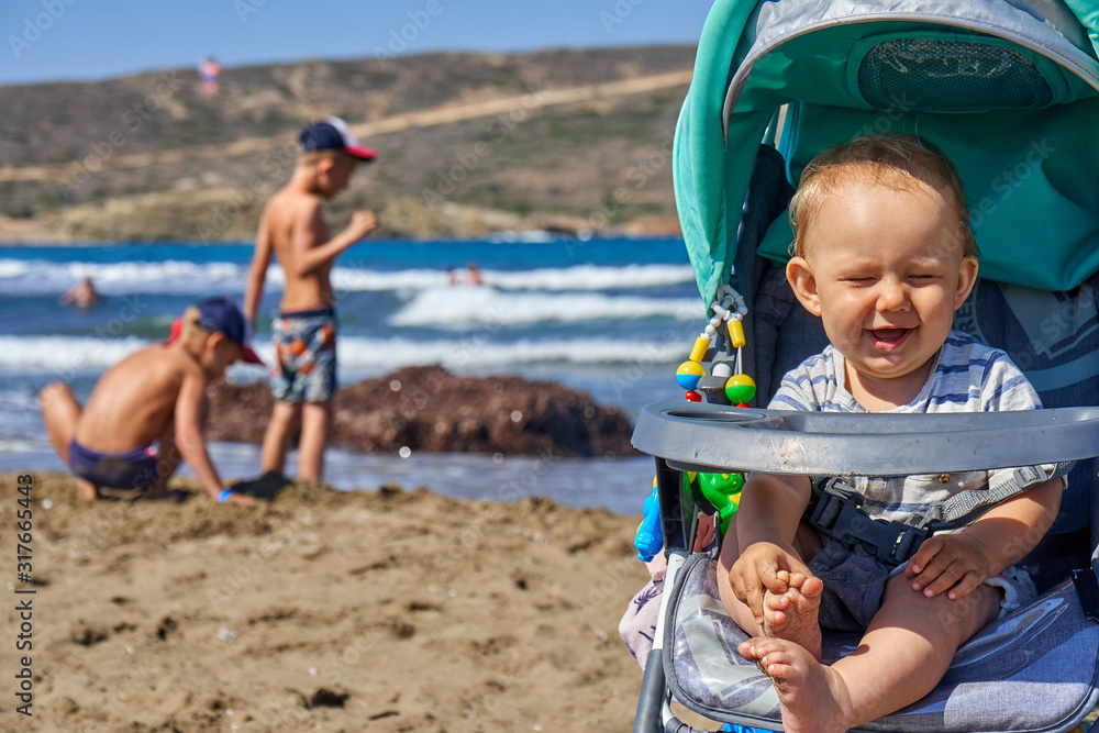 Fototapeta premium Joyful baby boy in stroller on beach in summer