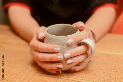 Closeup of womans hands holding a mug or cup of drink over light wooden table.