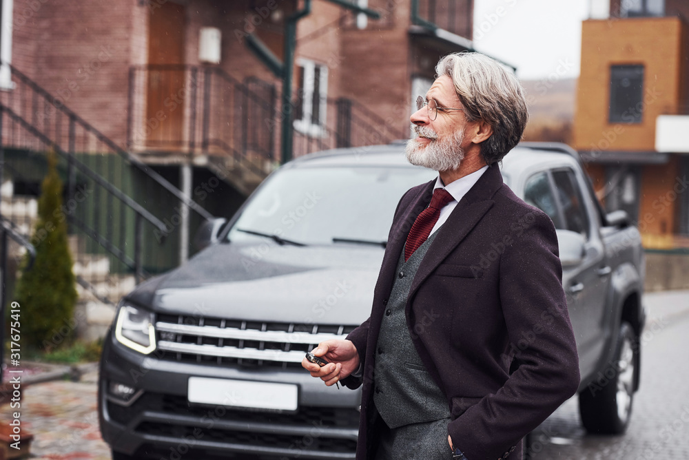 Fashionable senior man with gray hair and beard is outdoors on the street near his car Fashionable senior man with gray hair and beard is outdoors on the street near his car
