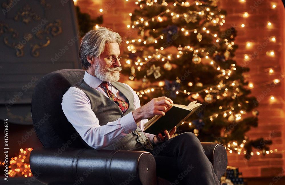 Portrait of stylish senior with grey hair and beard reading book in decorated christmas room
