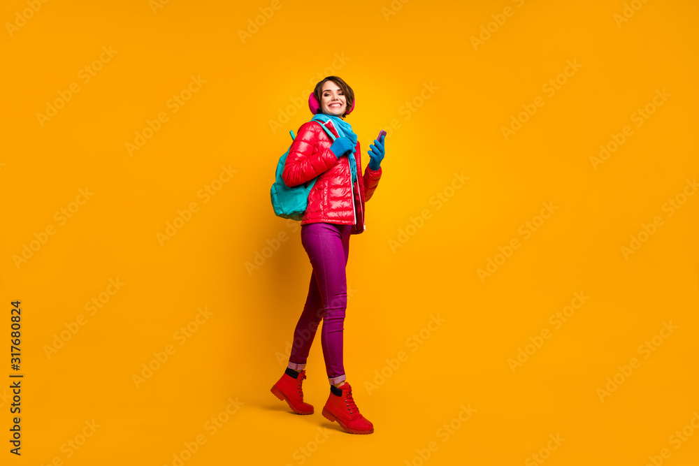 Full body profile photo of pretty lady walking down street using telephone online maps wear casual red coat boots blue scarf gloves ear covers pants isolated yellow color background