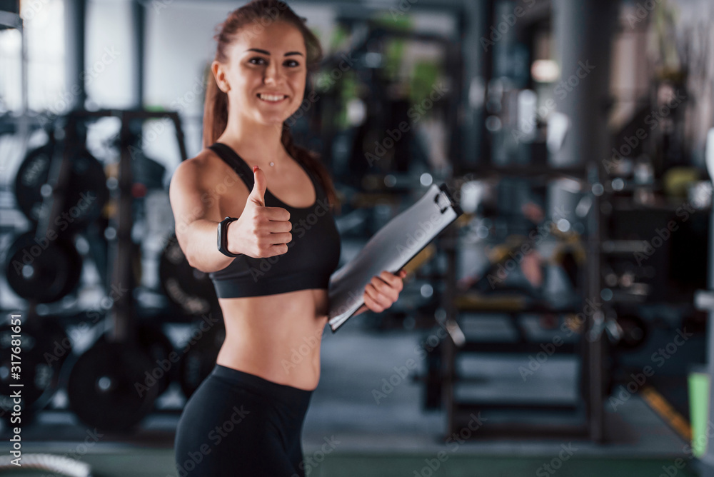 Young female fitness personal trainer with notepad standing in the gym with thumb up Young female fitness personal trainer with notepad standing in the gym with thumb up