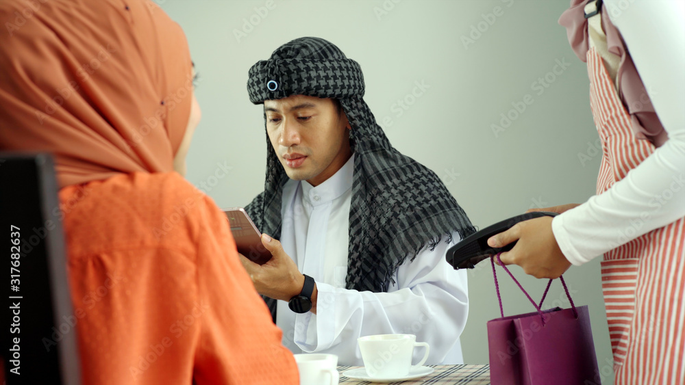 Young beautiful Asian Muslim man and women enjoying a relaxing moment in the coffee shop on a bright sunny day using mobile phone, smartwatch for mobile payment and QR code scan