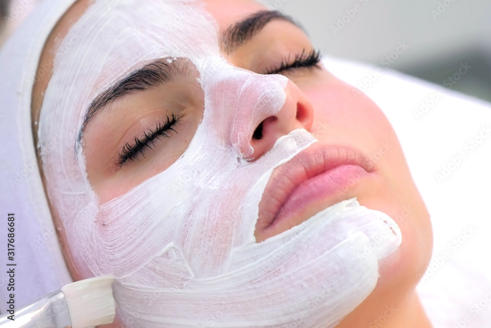 Cosmetologist is applying white mask on woman client face in beauty clinic. Portrait of woman closeup. Beautician making beauty facial skincare procedure to patient. Beauty industry concept.