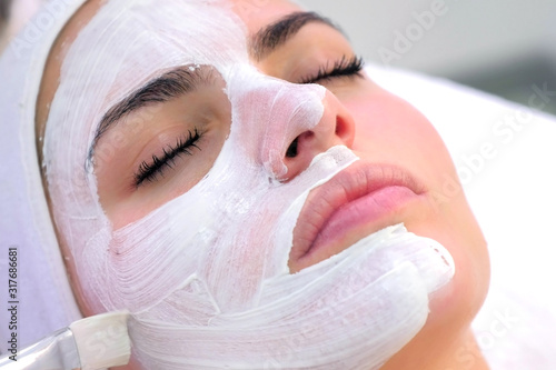 Cosmetologist is applying white mask on woman client face in beauty clinic. Portrait of woman closeup. Beautician making beauty facial skincare procedure to patient. Beauty industry concept.