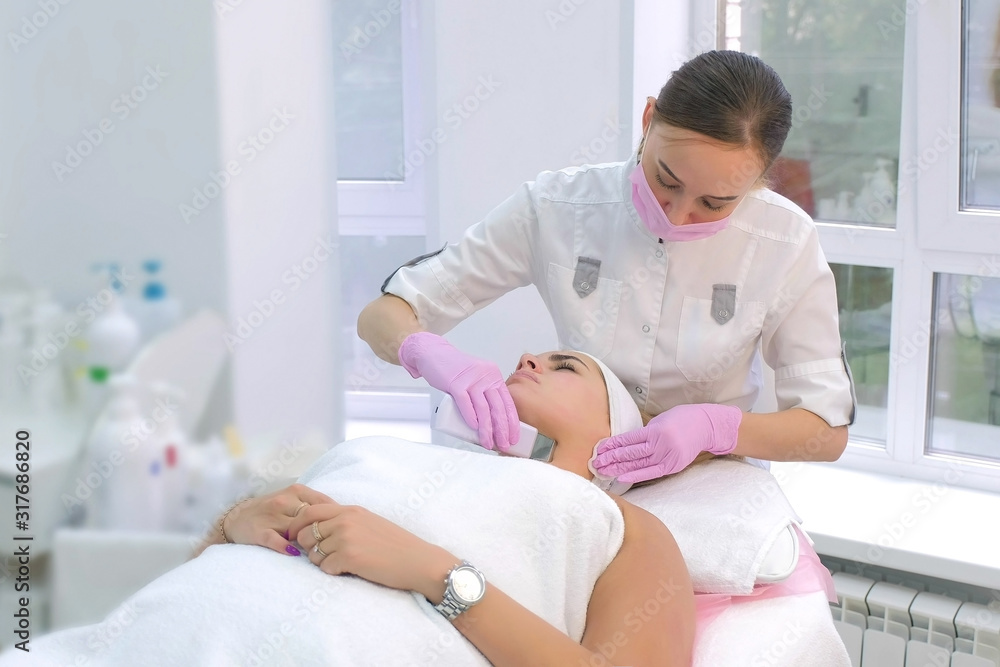 Cosmetologist making ultrasonic face cleaning procedure for young woman client in clinic. Beutician wiping face cotton pads. Beauty procedure in cosmetology salon. Beauty industry concept. Cosmetologist making ultrasonic face cleaning procedure for young woman client in clinic. Beutician wiping face cotton pads. Beauty procedure in cosmetology salon. Beauty industry concept.