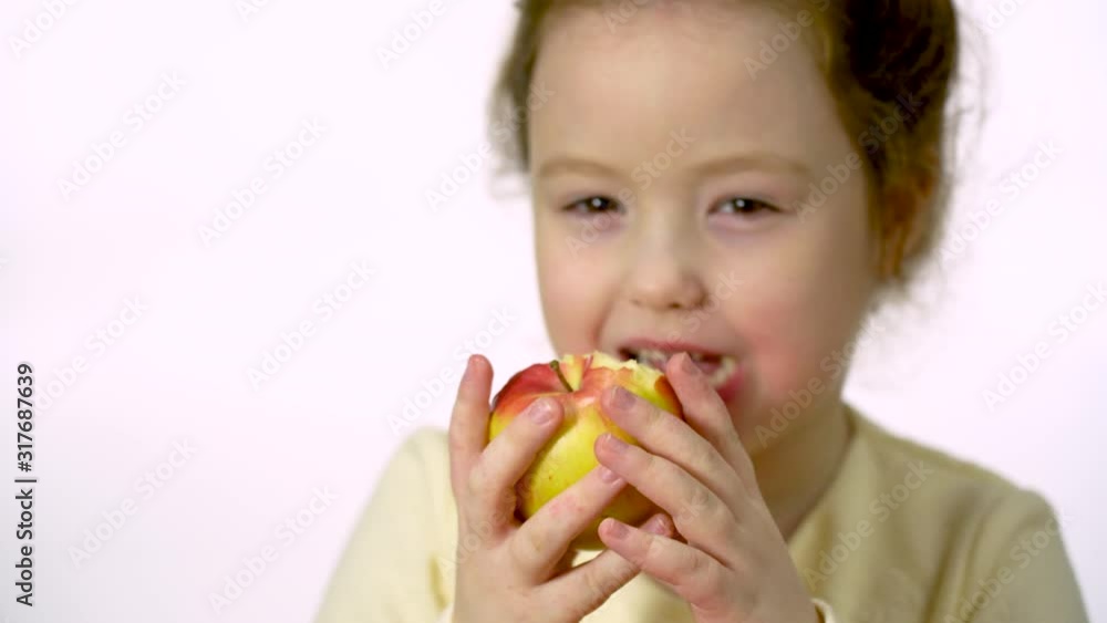 Girl 5-6 years old with long hair happily eats an apple in the studio on a white background