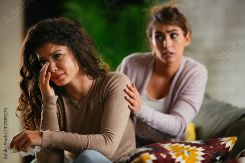 Woman broke up with boyfriend  she is crying and sister is trying to calm her down. Beautiful women sitting in living room. 