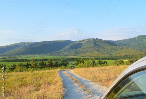 Stony country road on which there is a car. Ahead are vineyards and hills with greenery.
