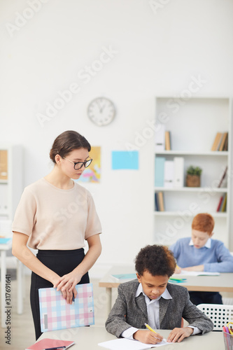 Young woman as a teacher watching for her student who working at his desk during a lesson