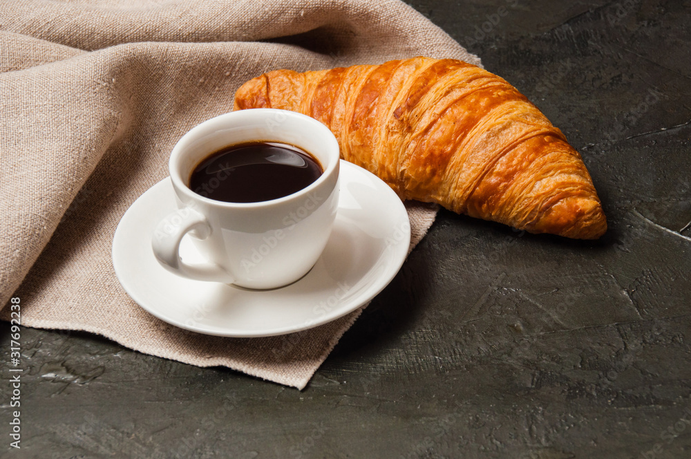 Croissant and cup of coffee and saucer on dark background with a gray linen napkin Croissant and cup of coffee and saucer on dark background with a gray linen napkin