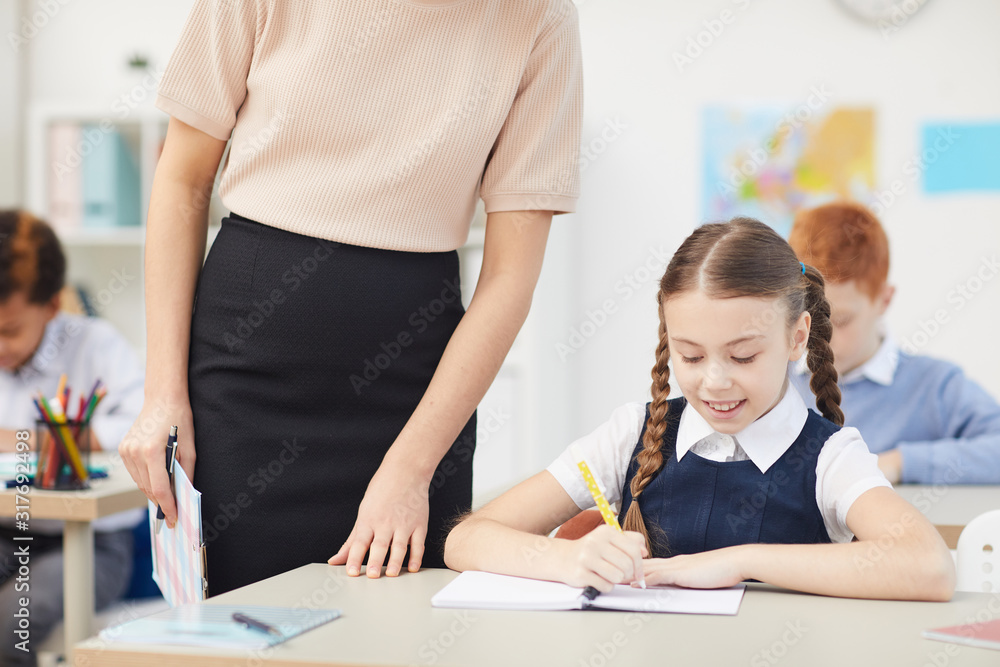 Smiling schoolgirl sitting at desk writing in notebook while teacher standing and controlling her work in the classroom Smiling schoolgirl sitting at desk writing in notebook while teacher standing and controlling her work in the classroom