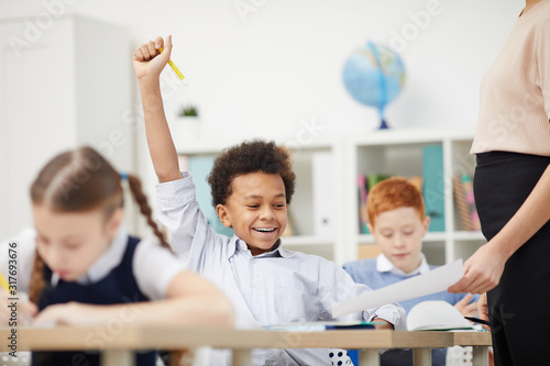 African schoolboy sitting at his desk and happy with good mark while teacher showing him the paper in the classroom