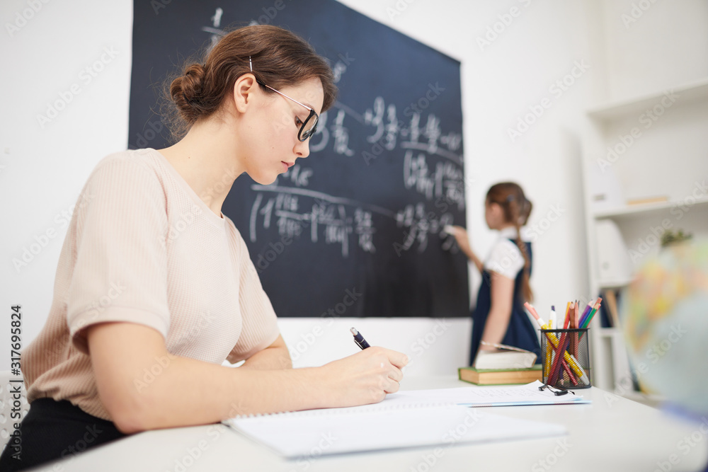 Fototapeta premium Young teacher sitting at the table and writing while schoolgirl working at the blackboard in the classroom at school