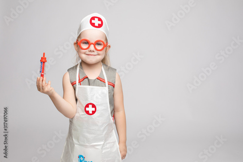 Canvas Print Little cute funny girl playing wearing doctor uniform holding toy syringe looking at camera