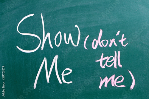 The phrase Show Me, Don't Tell Me written by hand in white chalk on a used blackboard. Often used in business but more so as a guide for novel and book writers
