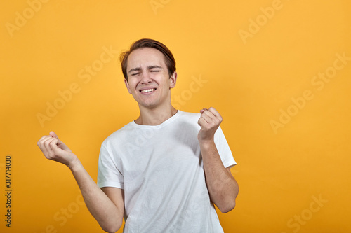 Cute guy clenched hands into fists and raised them, closing his eyes rejoicing in white t-shirt on yellow background
